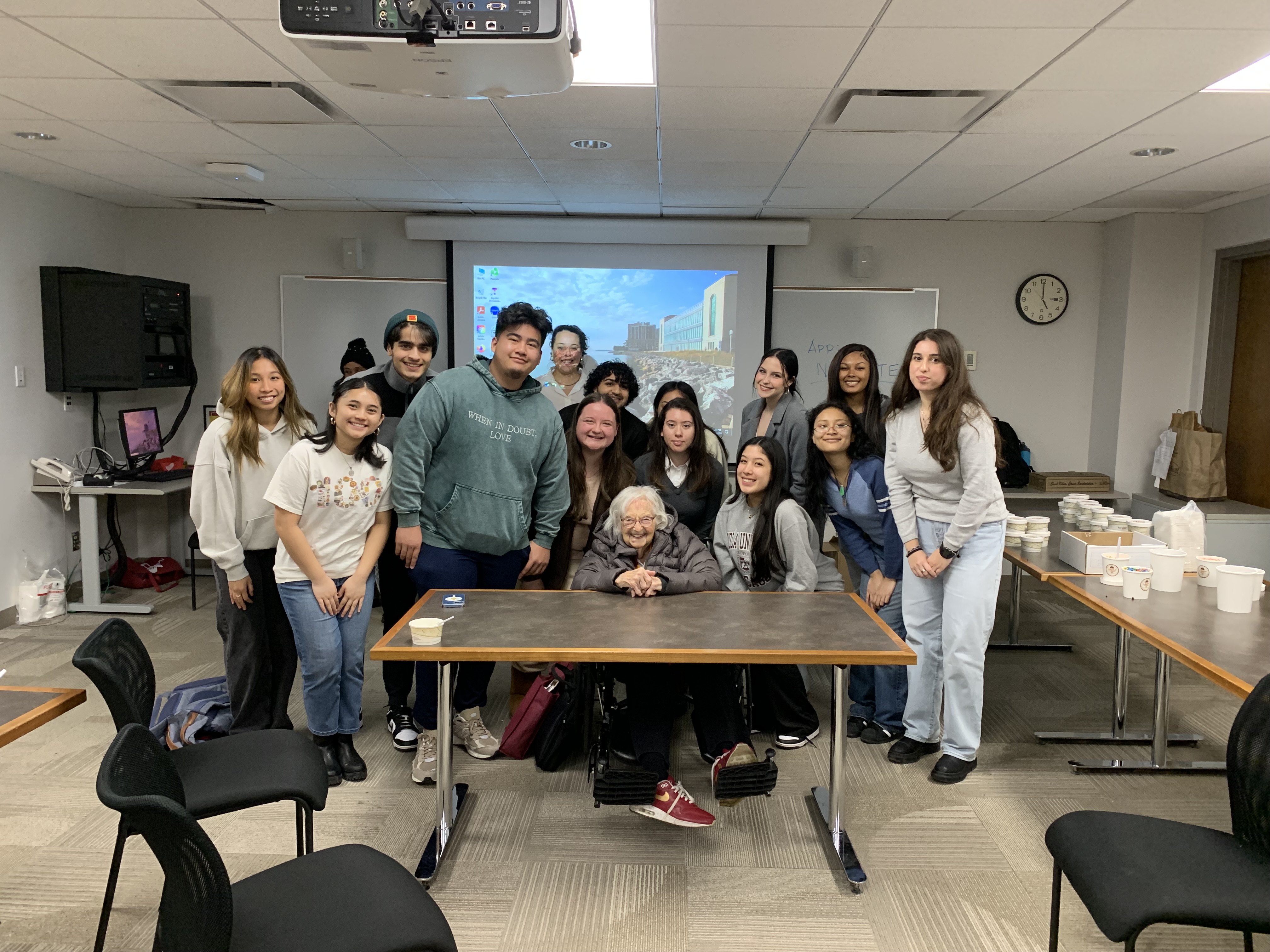 A group of Loyola University Chicago Leadership Scholars Program students with the late Sister Jean. Sister Jean is seated at a table, wearing her iconic Nike shoes, while the group leans in and stands around her in a classroom, smiling.